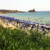 Spiaggia affollata con ombrelloni blu e un castello visibile in lontananza nel mare.