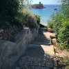 Sentiero scende verso la spiaggia, con un castello su un'isola in lontananza.