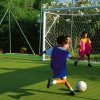 Bambini che giocano a calcio su un campo verde con porta e reti protettive.