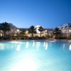 Piscina illuminata al crepuscolo, con palme e edificio sullo sfondo.