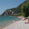 Spiaggia di ciottoli con bagnanti e montagne, accanto ad alberi verdi.