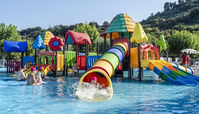 Bambini giocano in piscina con scivoli colorati.