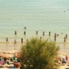 Persone in acqua poco profonda su una spiaggia con ombrelloni colorati e vegetazione.