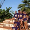 Gruppo sorridente con t-shirt viola seduto su gradini tra piante tropicali.
