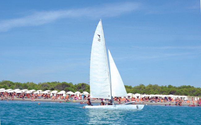 Vela bianca sul mare con spiaggia e ombrelloni sullo sfondo.