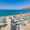 Spiaggia sabbiosa con ombrelloni blu e gialli, mare cristallino e colline in lontananza.