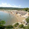 Spiaggia con ombrelloni e lettini, persone sul bagnasciuga, mare e vegetazione sullo sfondo.