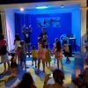 Bambini che ballano su un palco illuminato di blu con un piccolo pubblico seduto intorno.